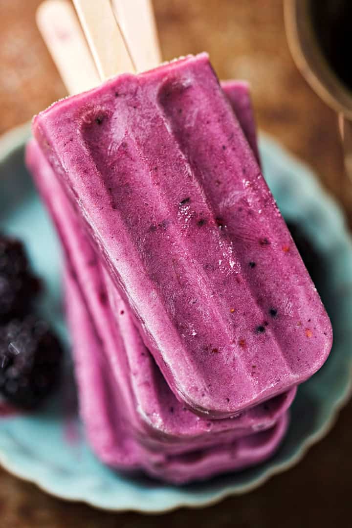 Three purple blackberry Greek yogurt popsicles stacked on a turquoise plate with two blackberries beside them, shown from above.