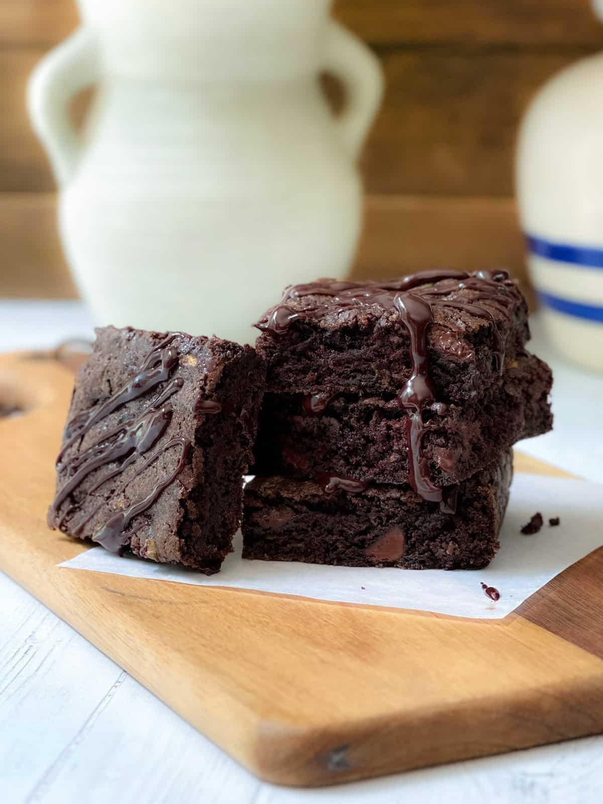 Three thick zucchini brownies stacked on a wooden board, drizzled with chocolate sauce, with ceramic vases in the background.