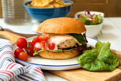 Side view of Firecracker Chicken Burgers on a white and blue plate with lettuce and tomatoes on a wood cutting board