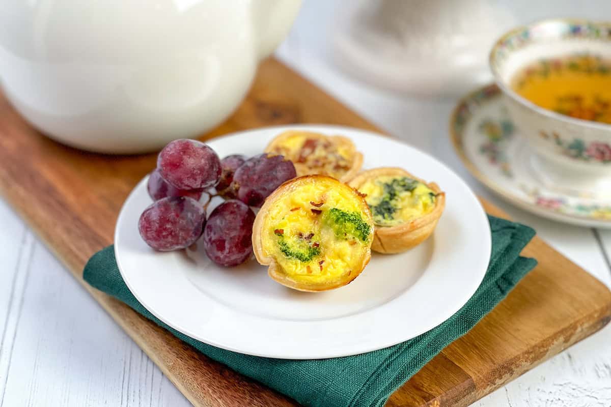A white plate with three mini quiches and a cluster of red grapes sits on a wooden board next to a teacup and teapot.