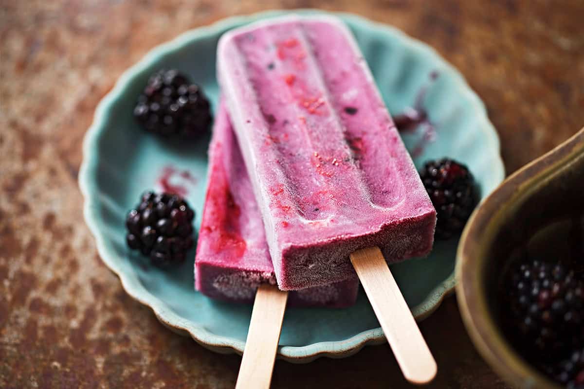 Two purple blackberry Greek yogurt popsicles rest on a teal plate, with fresh blackberries scattered around them.