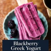 Three purple blackberry Greek yogurt popsicles on a plate with fresh blackberries, next to a ceramic jug. Text overlay reads: "Blackberry Greek Yogurt Popsicles (Naturally Sweetened)". These delicious blackberry Greek yogurt popsicles are the perfect refreshing treat, made with wholesome ingredients for a naturally sweet twist.