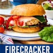 A plate featuring firecracker chicken burgers loaded with lettuce, melted cheese, and cherry tomatoes. Text: "Firecracker Chicken Burgers - Easy 4th of July Grilling Recipe.