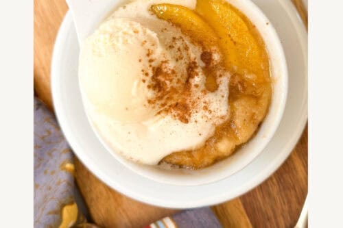 A white mug filled with peach mug cake, topped with vanilla ice cream and cinnamon, sits on a wooden board beside fresh peaches and a patterned cloth. Text reads: "3 Minute Peach Mug Cake. Peach cobbler in minutes!.