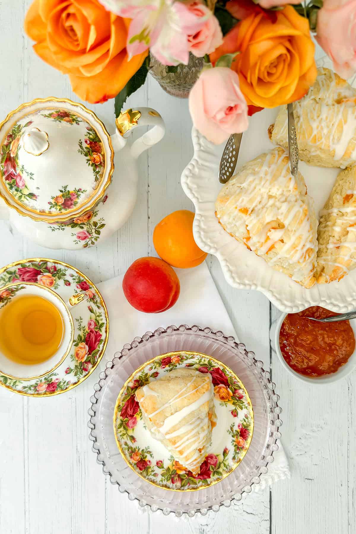 A floral tea set with a teapot, teacup filled with tea, apricot scones topped with icing and jam.