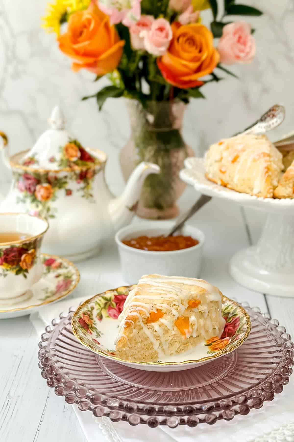 An apricot scone with white icing on a floral plate, accompanied by tea, a teapot, jam, and a vase of roses.