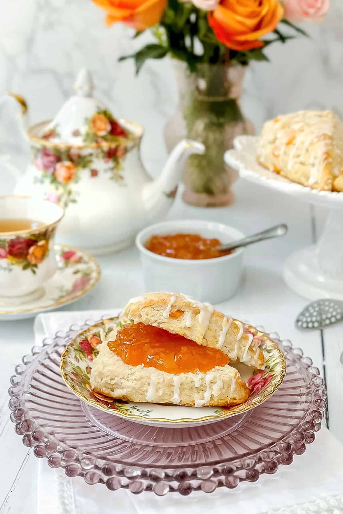 An Apricot Scone with icing and orange jam sits on a floral plate, surrounded by a teapot, tea cup, jam bowl, and a vase of orange roses in the background.