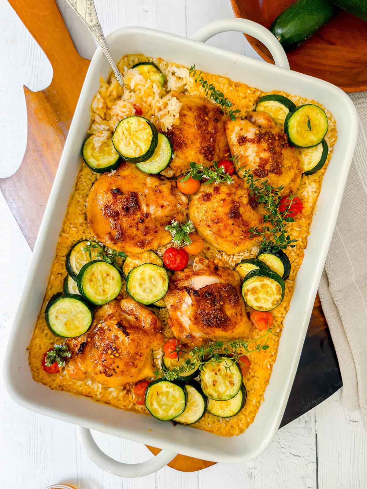 Baked chicken and rice served on a bed of fluffy grains, garnished with sliced zucchini, cherry tomatoes, and fresh herbs in a white casserole dish.