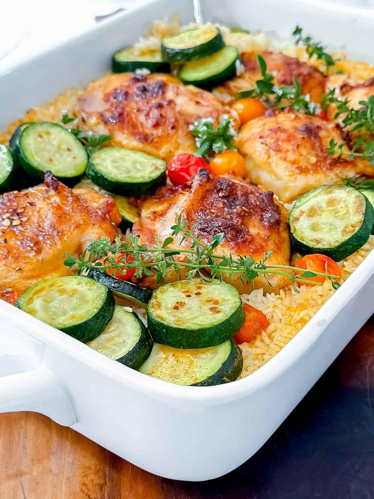 Baked chicken and rice with zucchini slices, cherry tomatoes, and fresh thyme in a white casserole dish.