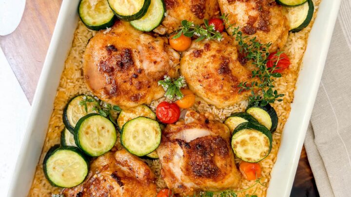 A white baking dish filled with baked chicken and rice, sliced zucchini, cherry tomatoes, and fresh herbs makes for a colorful and wholesome meal.
