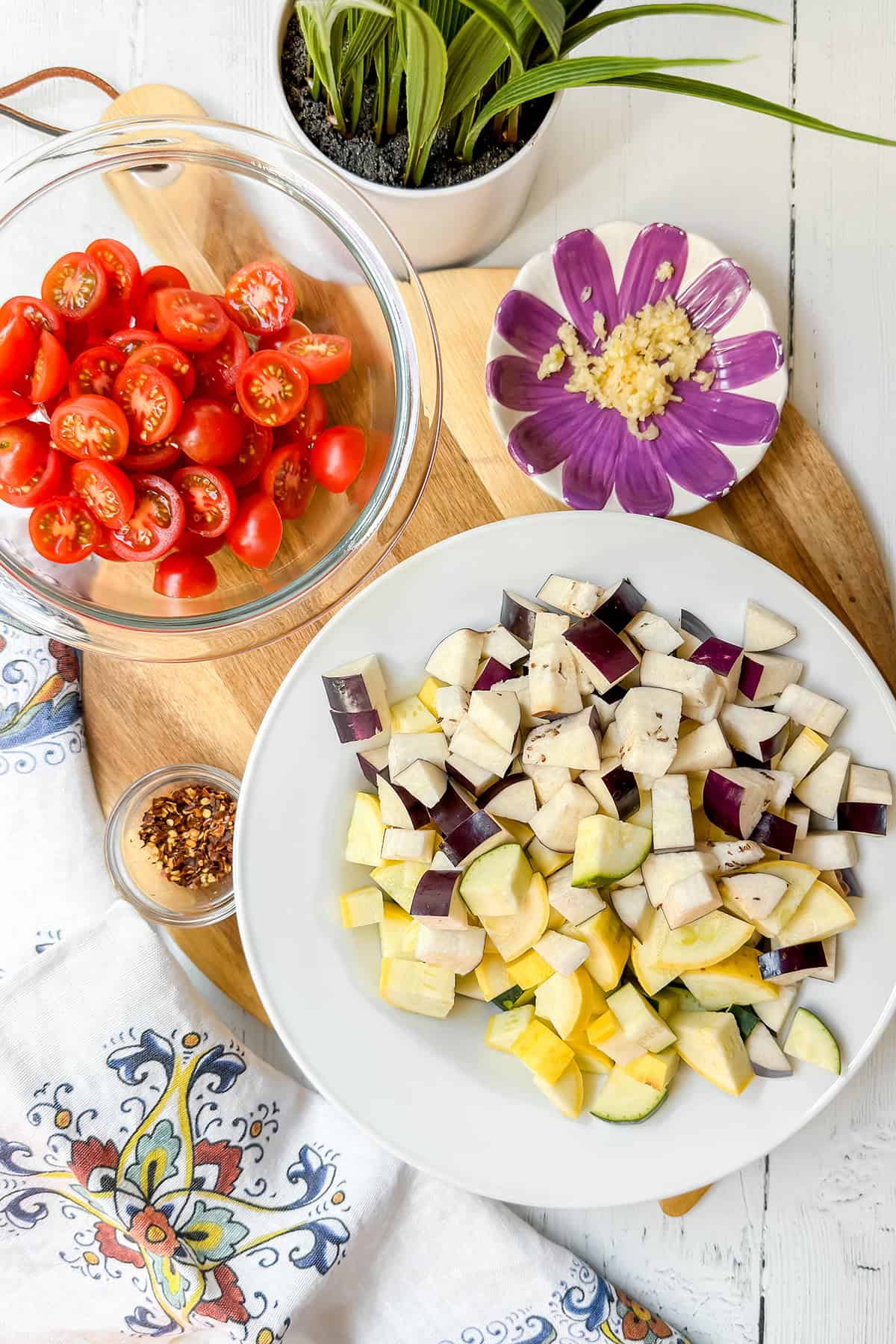 A bowl of halved cherry tomatoes, a plate of diced eggplant and squash, minced garlic on a floral dish, and a small bowl of red pepper flakes await to become the perfect garden pasta on a wooden board.