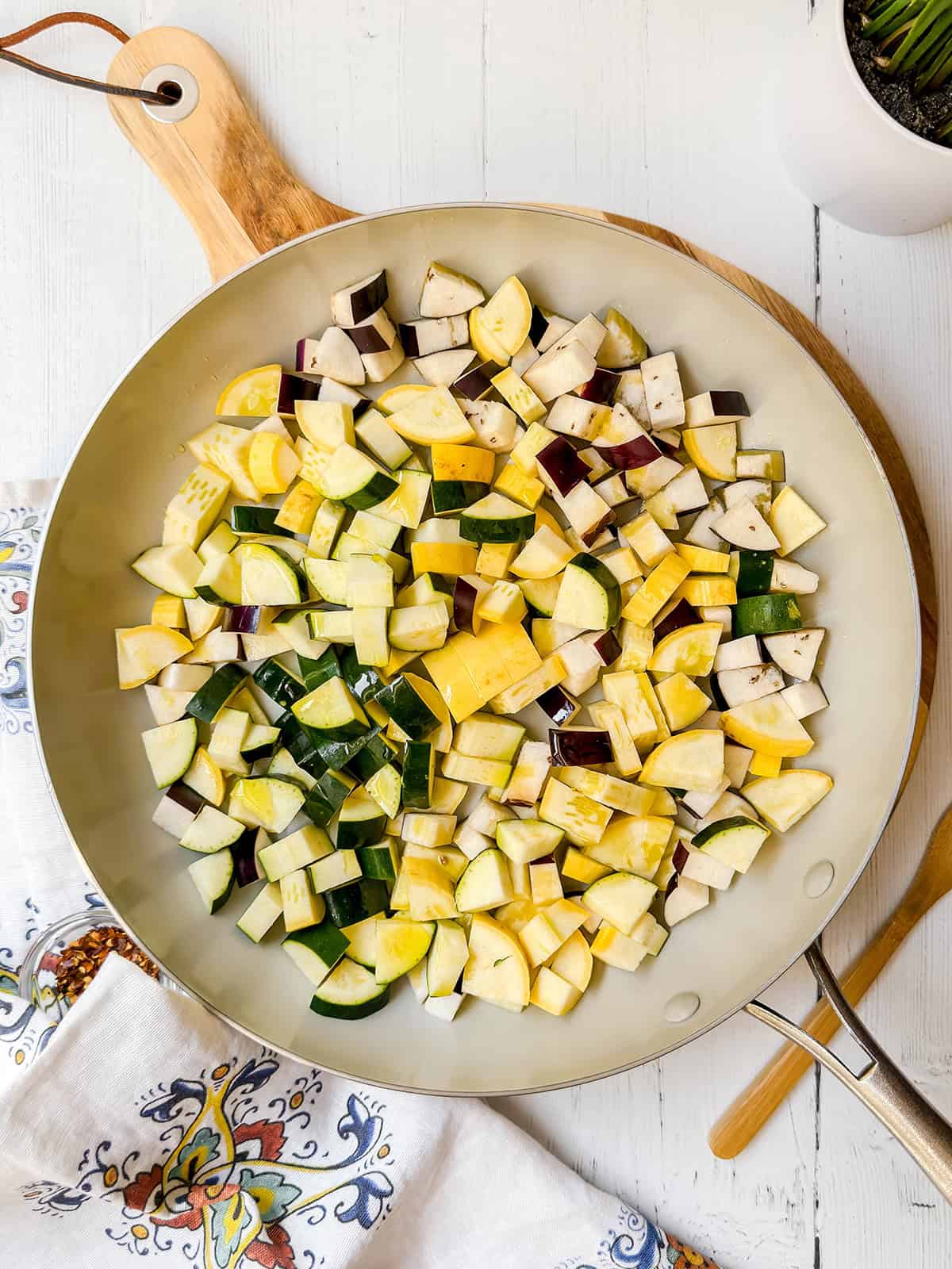 A large skillet filled with chopped yellow squash, zucchini, and eggplant for a fresh garden pasta sits on a white wooden surface next to a patterned towel and wooden utensils.