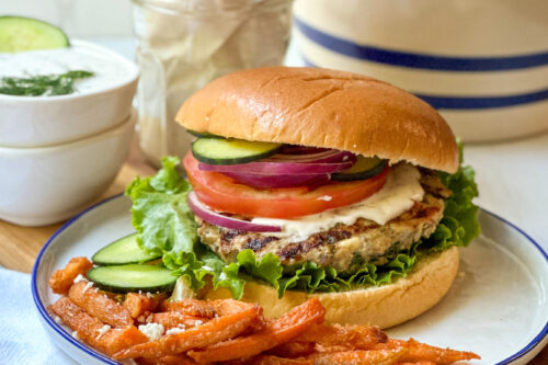 Side view of grilled Greek Turkey Burger on a bun with cucumber slices, tzatziki sauce, tomato and red onion and served with sweet potato fries