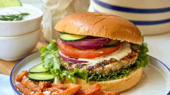 Side view of grilled Greek Turkey Burger on a bun with cucumber slices, tzatziki sauce, tomato and red onion and served with sweet potato fries