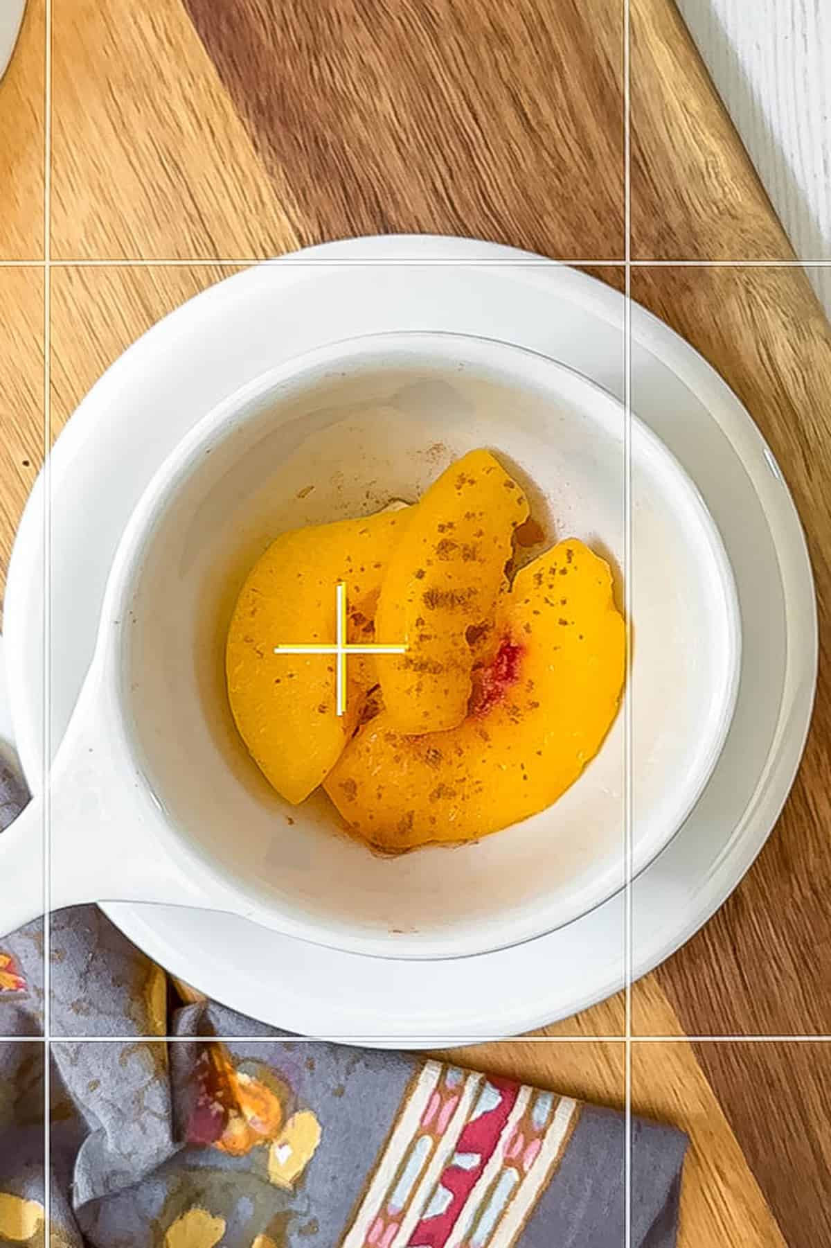 A white bowl with sliced peaches sprinkled with cinnamon sits on a white plate atop a wooden surface, next to a patterned napkin-perfect inspiration for your next peach mug cake.