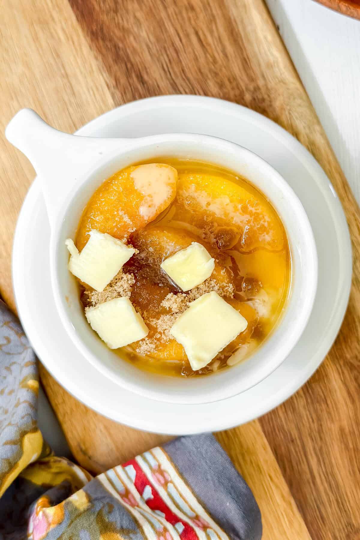 A white bowl filled with peach slices, perfect for a peach mug cake, is topped with cubes of butter and sprinkled with brown sugar. It sits on a matching plate on a wooden surface, with a patterned cloth partially visible nearby.