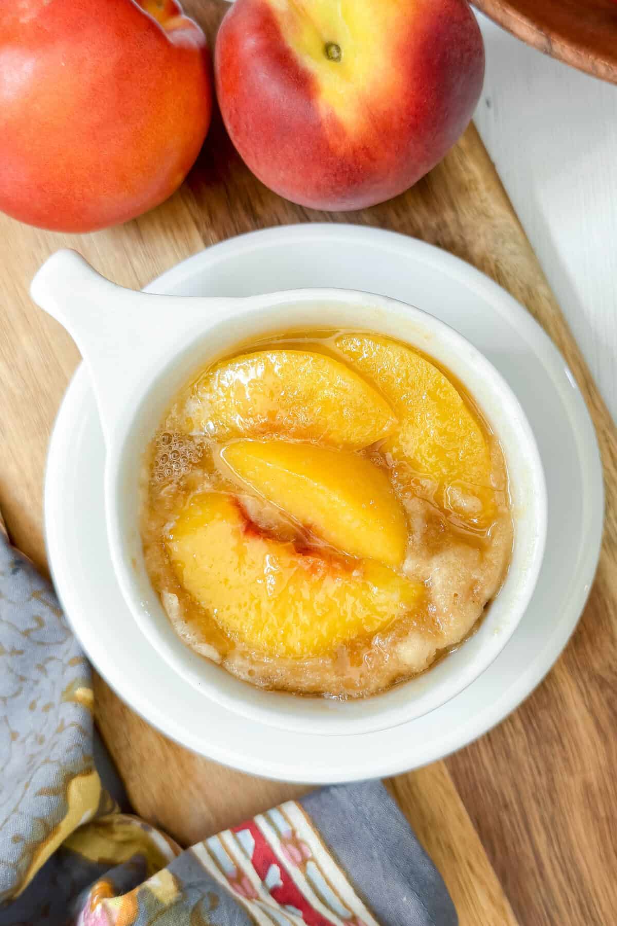 A mug of peach cobbler with golden peach slices sits on a white plate, next to fresh peaches and a colorful cloth&mdash;perfect inspiration for making an easy peach mug cake at home.