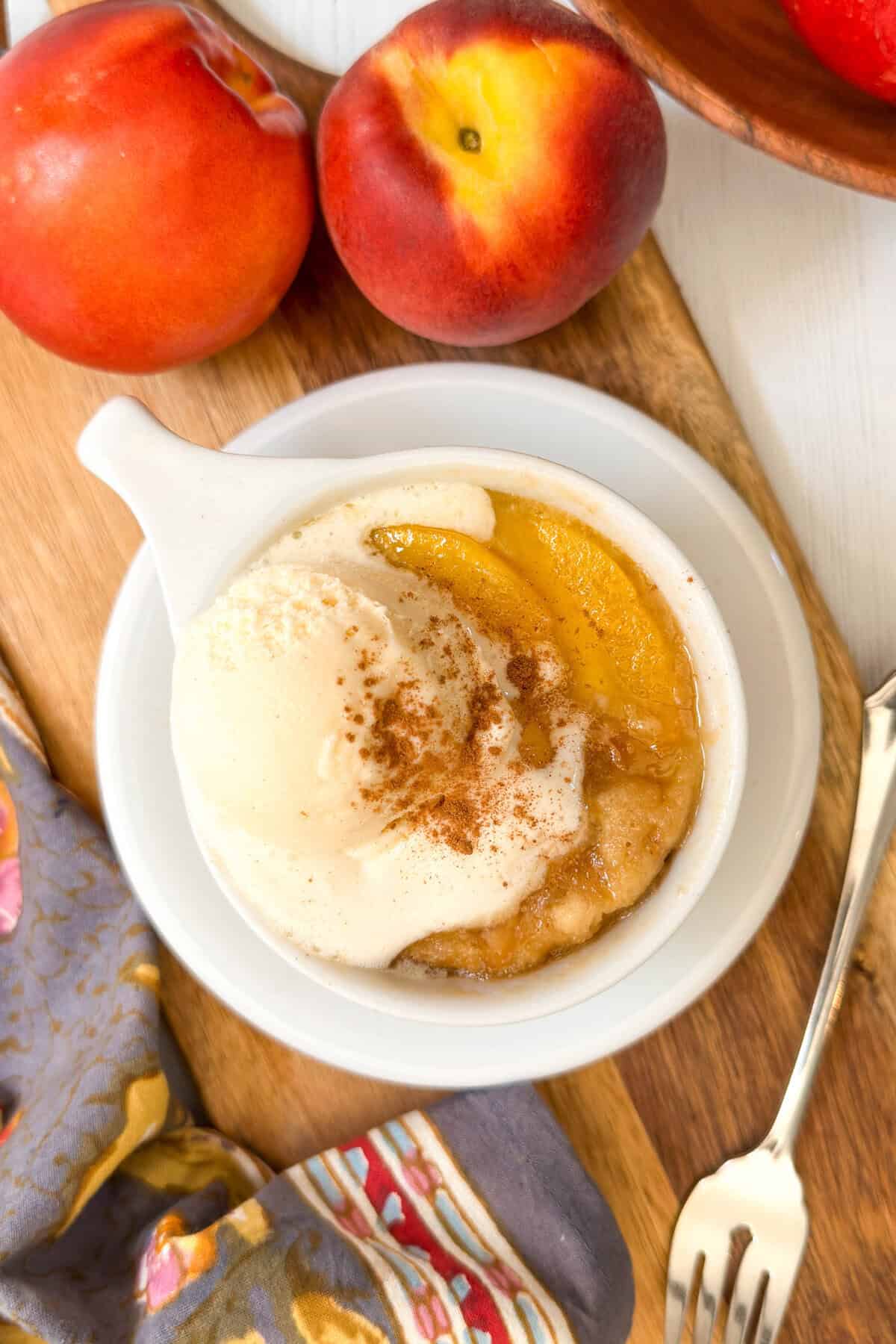 A bowl of peach cobbler topped with vanilla ice cream and sprinkled with cinnamon, placed on a wooden board next to fresh peaches, a fork, and a patterned cloth napkin&mdash;perfect for fans of classic treats or a quick peach mug cake.