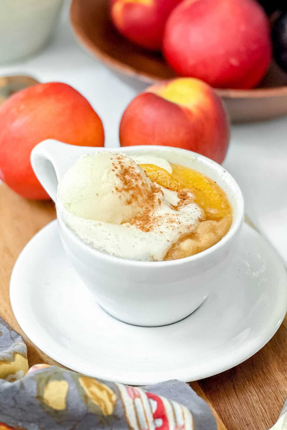 A white cup of peach mug cake topped with vanilla ice cream and a sprinkle of cinnamon, served on a saucer, with fresh peaches and a wooden bowl in the background.