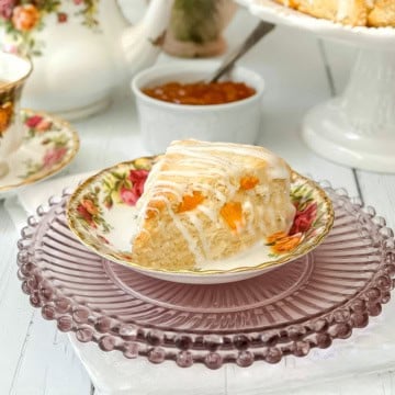 A slice of glazed Apricot Scone with rests on a tea plate. A bowl of jam and a teapot are in the background.