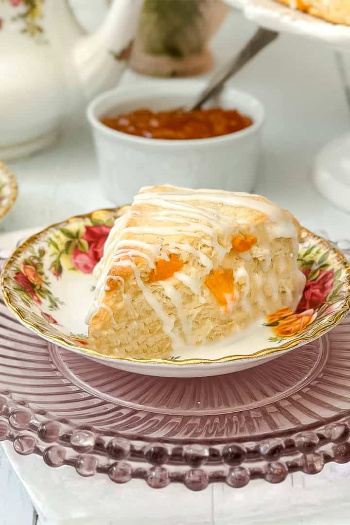 A slice of glazed Apricot Scone with rests on a tea plate. A bowl of jam and a teapot are in the background.