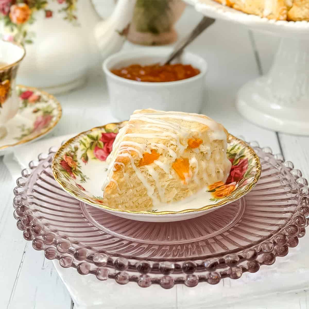A slice of glazed Apricot Scone with rests on a tea plate. A bowl of jam and a teapot are in the background.