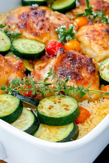 Baked chicken and rice with zucchini slices, cherry tomatoes, and fresh thyme in a white casserole dish.