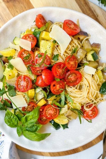 A plate of garden pasta topped with cherry tomatoes, diced zucchini, eggplant, fresh basil, parsley, and shaved parmesan cheese on a white plate.