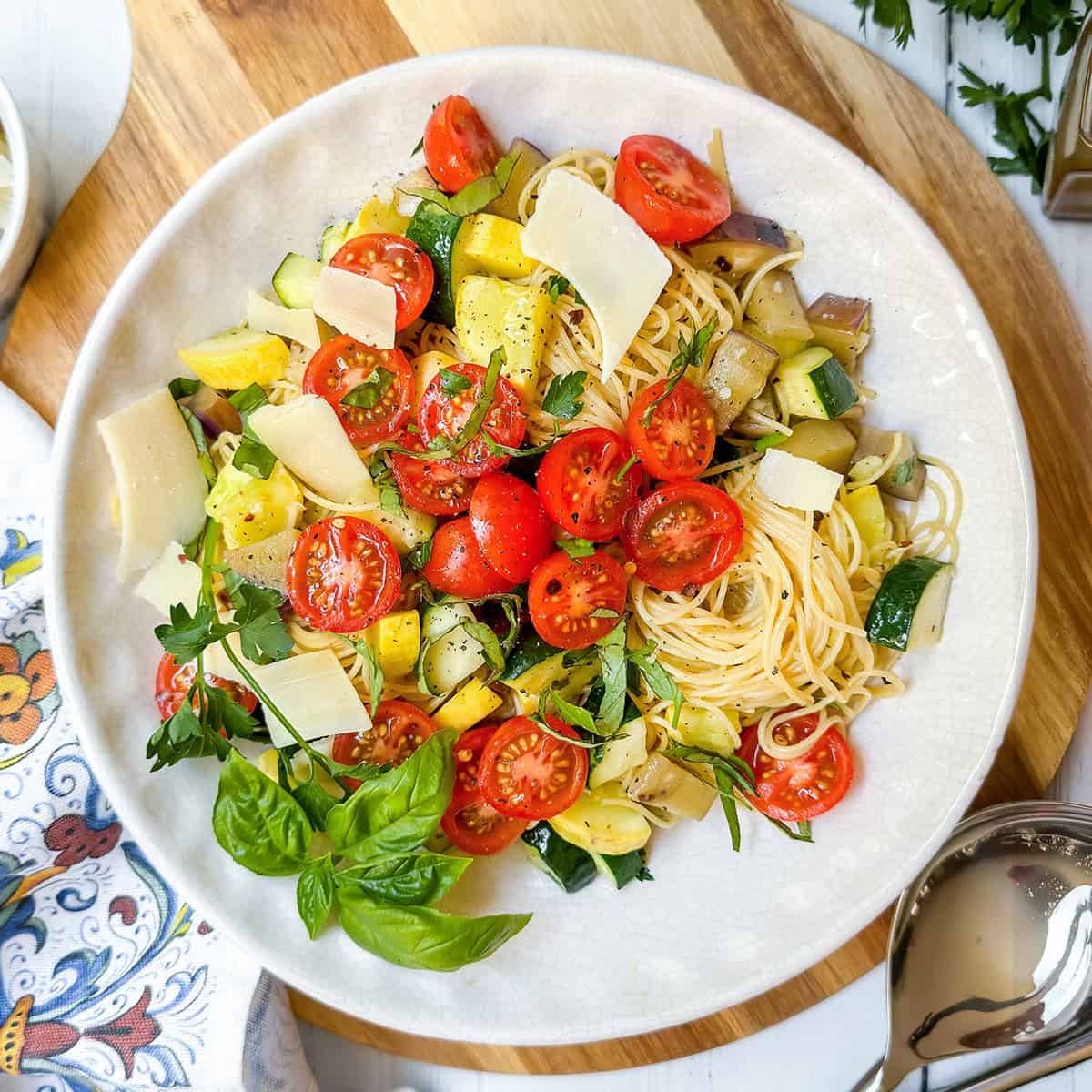 A plate of garden pasta topped with cherry tomatoes, diced zucchini, eggplant, fresh basil, parsley, and shaved parmesan cheese on a white plate.
