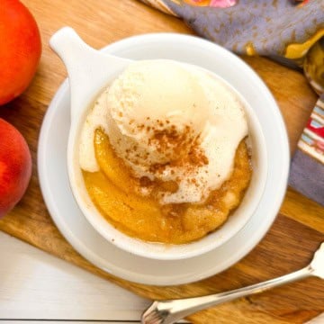 A peach mug cake topped with vanilla ice cream and cinnamon sits on a wooden board next to peaches and a patterned cloth, evoking the cozy flavor of a homemade peach mug cake.