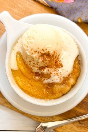 A peach mug cake topped with vanilla ice cream and cinnamon sits on a wooden board next to peaches and a patterned cloth, evoking the cozy flavor of a homemade peach mug cake.