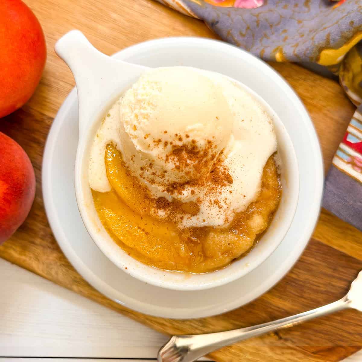 A peach mug cake topped with vanilla ice cream and cinnamon sits on a wooden board next to peaches and a patterned cloth, evoking the cozy flavor of a homemade peach mug cake.