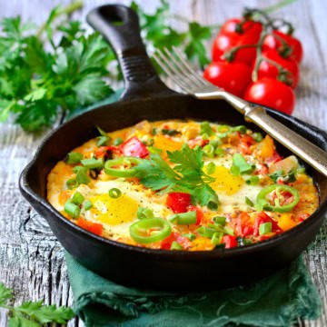 A cast iron skillet containing shakshuka with eggs, chopped vegetables, and herbs, garnished with fresh parsley and green onions; tomatoes and parsley are in the background.