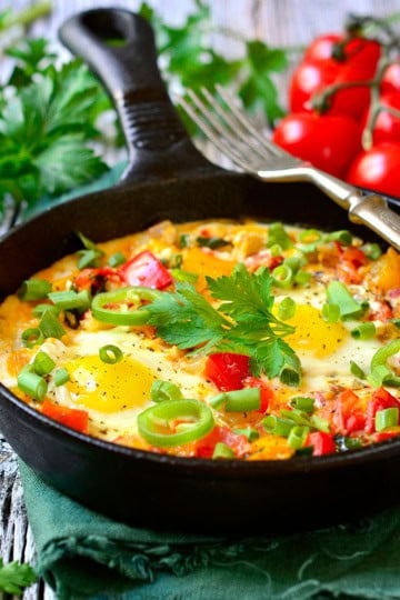 A cast iron skillet containing shakshuka with eggs, chopped vegetables, and herbs, garnished with fresh parsley and green onions; tomatoes and parsley are in the background.