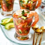 Glass filled with Mexican Shrimp Cocktail, topped with shrimp and parsley, served with avocado slices on the side and gold spoons nearby.