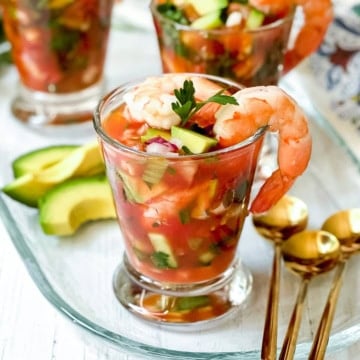 Glass filled with Mexican Shrimp Cocktail, topped with shrimp and parsley, served with avocado slices on the side and gold spoons nearby.