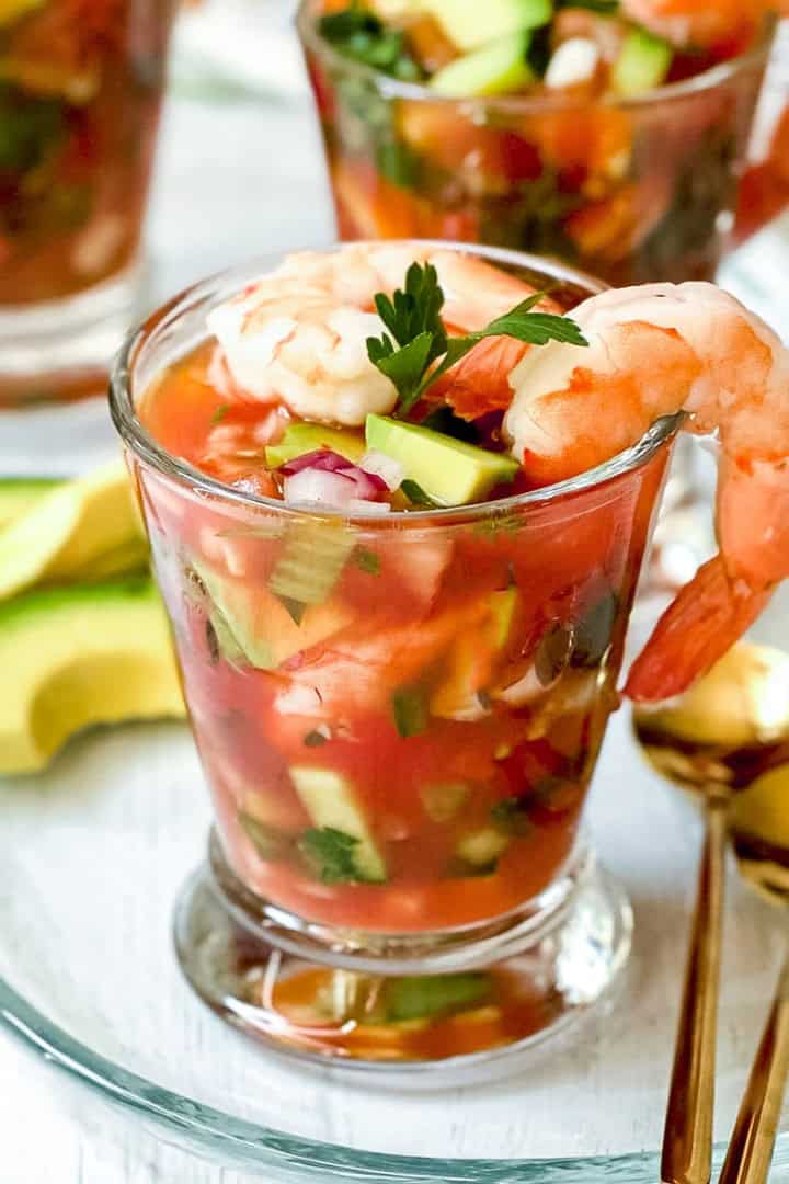 Glass filled with Mexican Shrimp Cocktail, topped with shrimp and parsley, served with avocado slices on the side and gold spoons nearby.