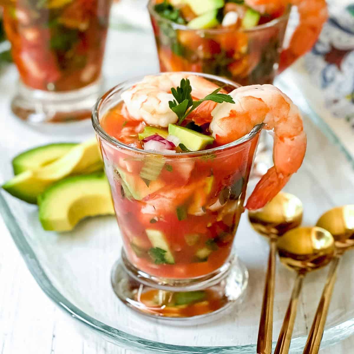 Glass filled with Mexican Shrimp Cocktail, topped with shrimp and parsley, served with avocado slices on the side and gold spoons nearby.