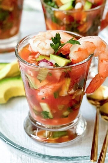 A glass filled with Mexican Shrimp Cocktail—plump shrimp, diced vegetables, and tangy tomato sauce—garnished with parsley, served alongside creamy avocado slices and elegant gold spoons.