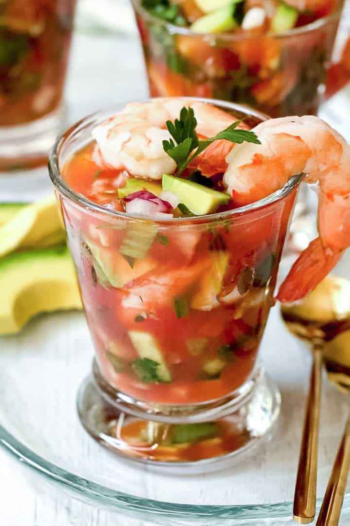 A glass filled with Mexican Shrimp Cocktail—plump shrimp, diced vegetables, and tangy tomato sauce—garnished with parsley, served alongside creamy avocado slices and elegant gold spoons.