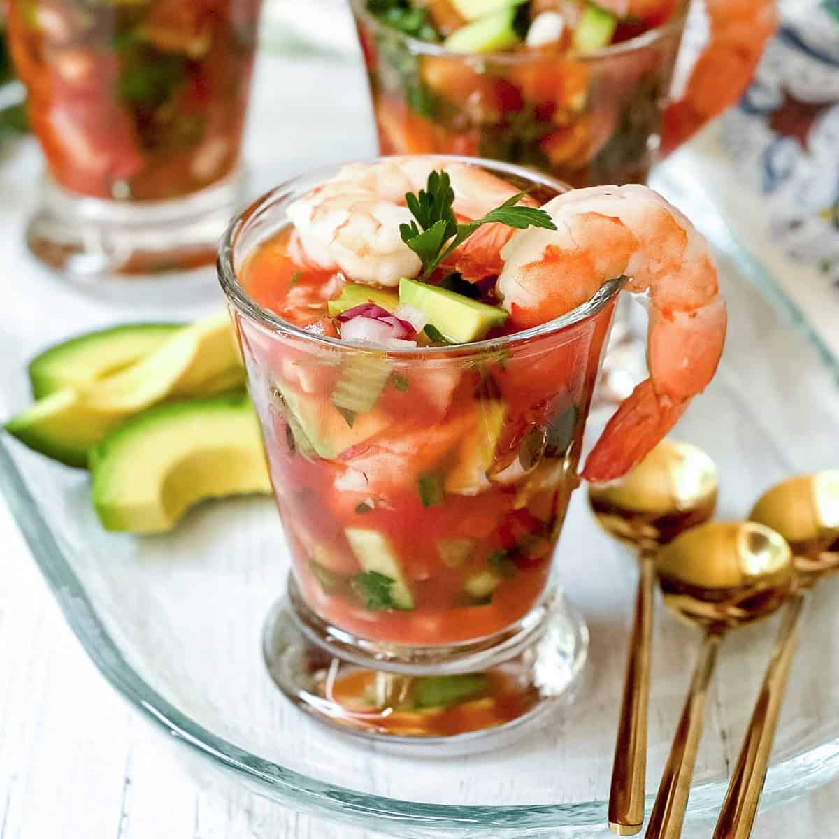 A glass filled with Mexican Shrimp Cocktail—plump shrimp, diced vegetables, and tangy tomato sauce—garnished with parsley, served alongside creamy avocado slices and elegant gold spoons.
