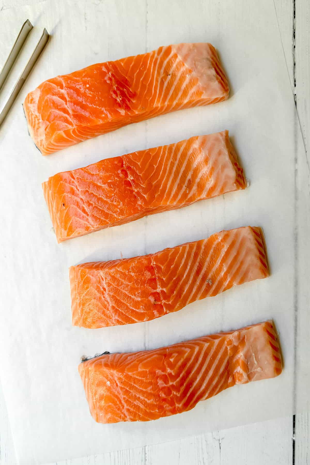 Four raw salmon fillets are arranged in a row on white parchment paper atop a white surface, with metal tongs placed nearby in the top left corner-perfect for your next salmon recipe.