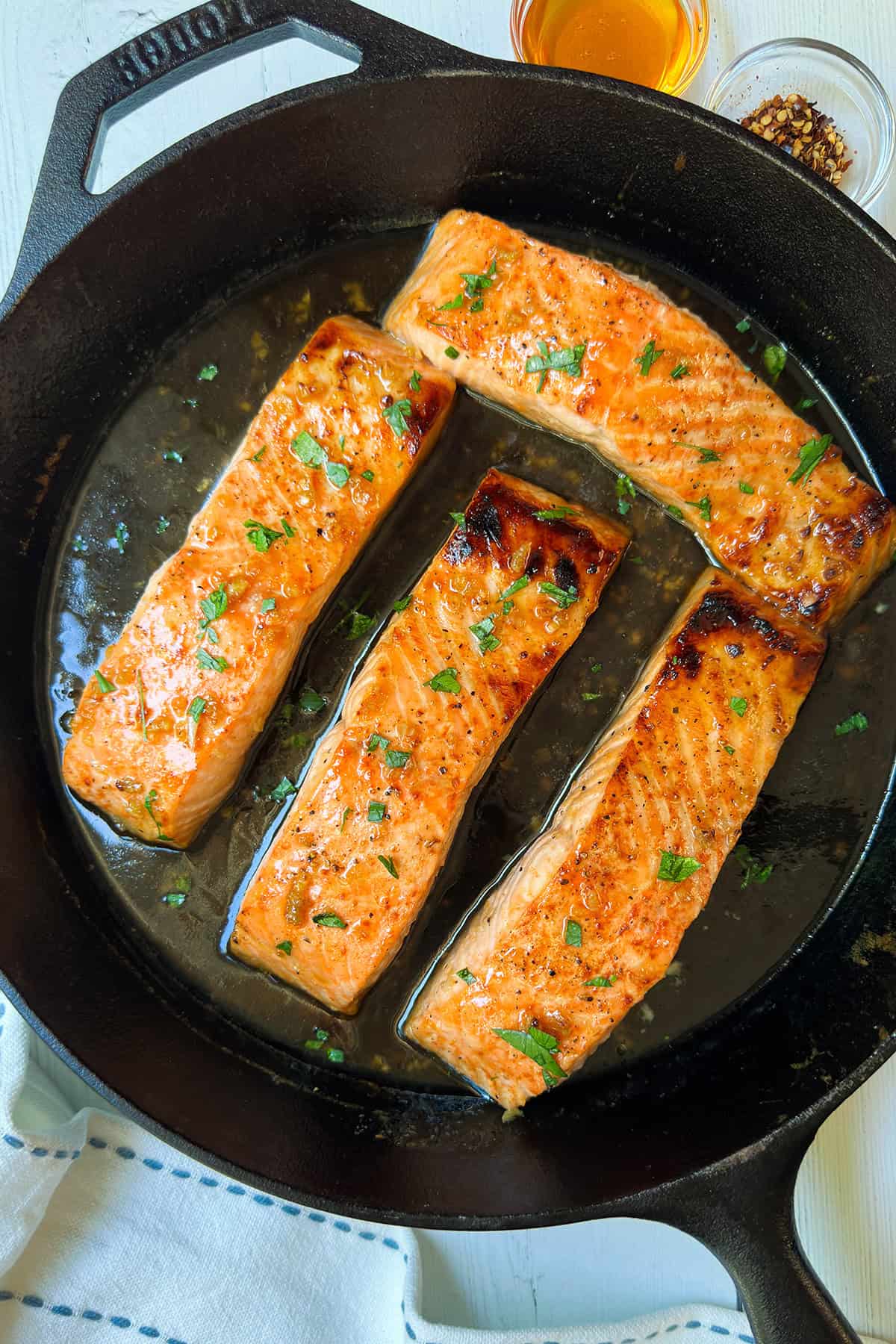 Four cooked salmon fillets garnished with herbs in a black cast iron skillet, with small bowls of sauce and seasonings nearby-perfect for your next salmon recipe.