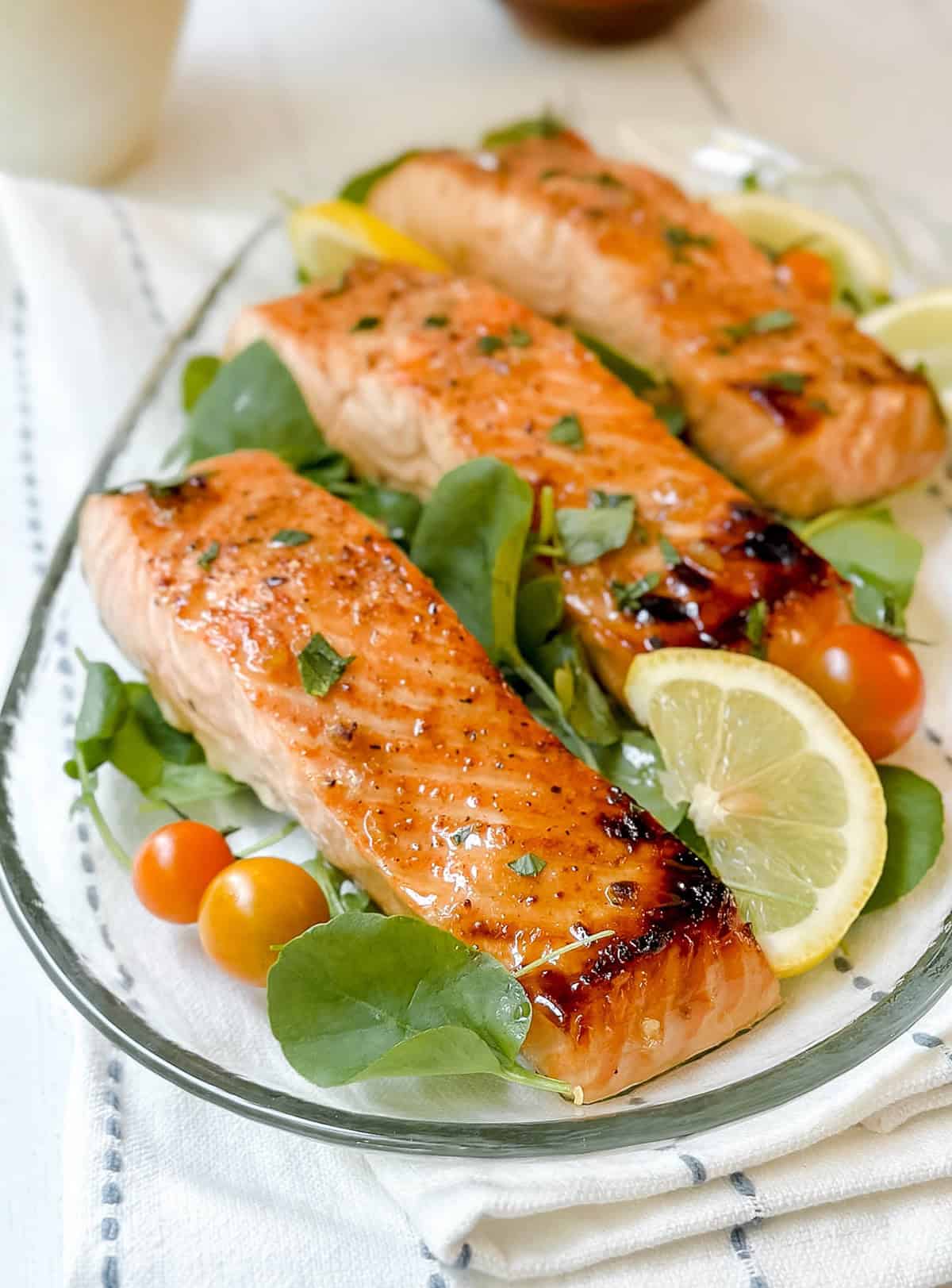 Three cooked salmon fillets on a bed of greens with cherry tomatoes and lemon slices, served on a clear glass plate-an easy yet elegant salmon recipe for any occasion.