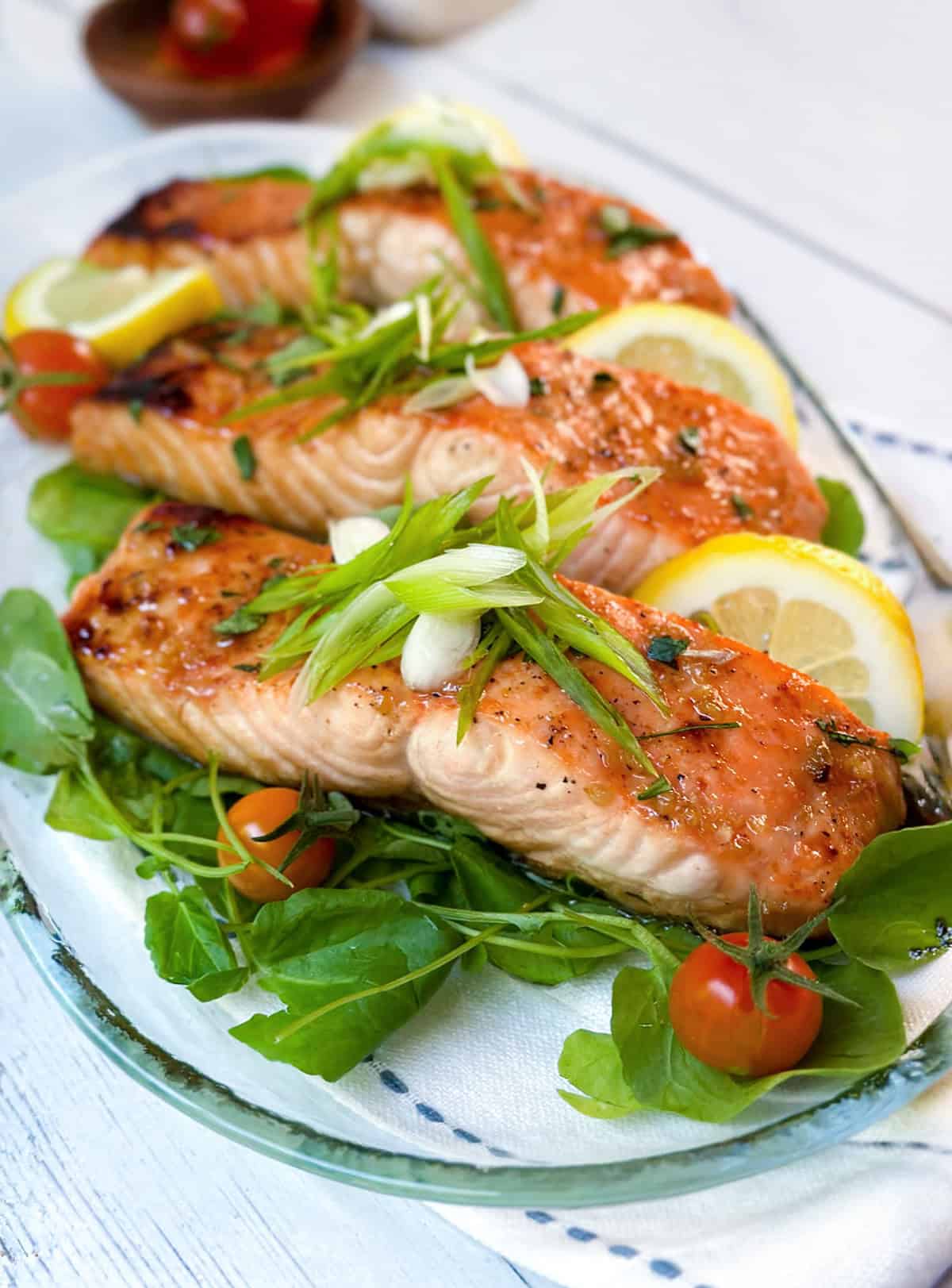 Three grilled salmon fillets served on a bed of greens with cherry tomatoes, lemon slices, and chopped green onions—a fresh and vibrant salmon recipe presented on a glass platter.