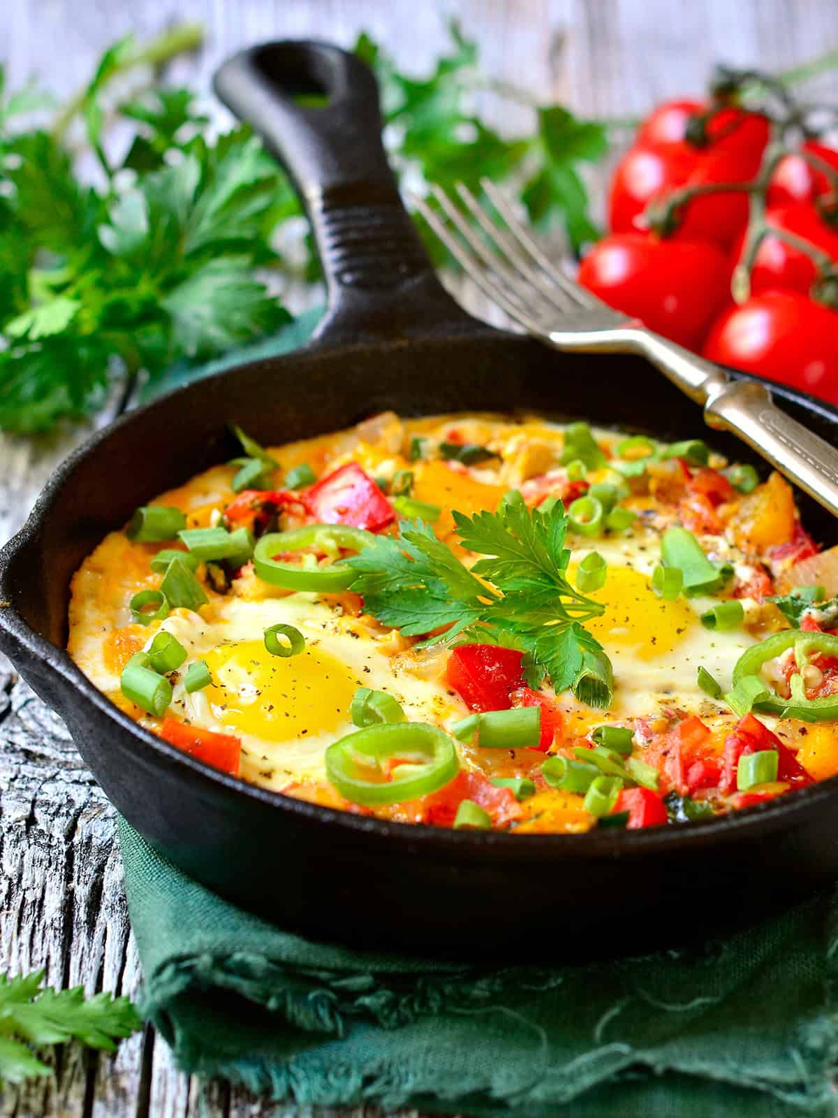 A cast iron skillet with Mediterranean shakshuka, topped with green onions and parsley, placed on a wooden surface with fresh tomatoes and herbs nearby.