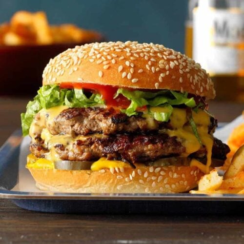 A double cheeseburger with lettuce, tomato, pickles, and melted cheese in a sesame seed bun, served with fries on a metal tray—perfect for your Labor Day menu ideas.