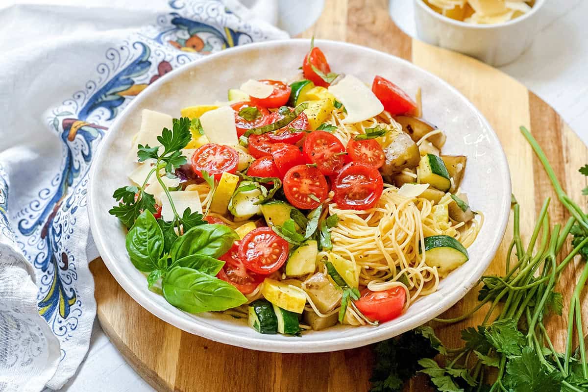 A bowl of garden pasta with cherry tomatoes, zucchini, yellow squash, fresh basil, parsley, and parmesan slices sits on a wooden board with herbs and a napkin nearby.
