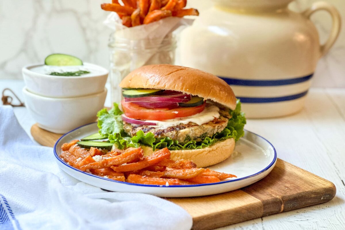 Side view of cooked Greek Turkey Burgers on a bun with tzatziki sauce, sliced cucumbers, tomatoes, and red onion.