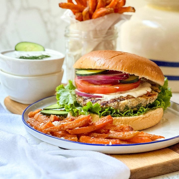 Side view of cooked Greek Turkey Burgers on a bun with tzatziki sauce, sliced cucumbers, tomatoes, and red onion.